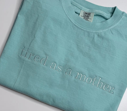 Teal t-shirt with 'tired as a mother' text on a light gray background