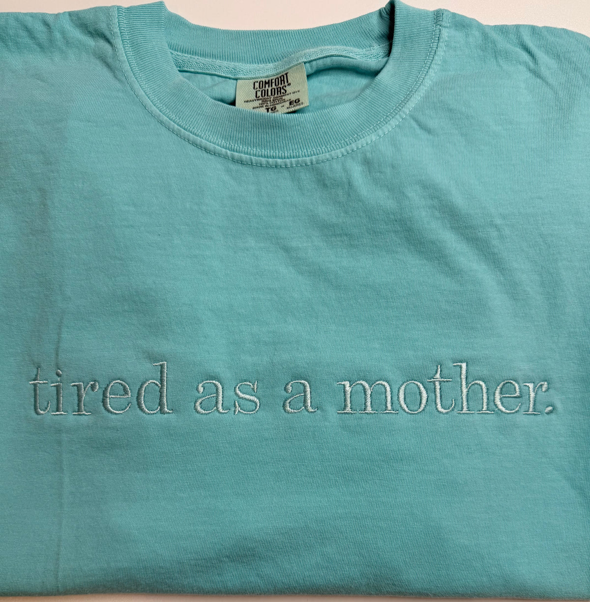 Teal t-shirt with 'tired as a mother' text on a beige background