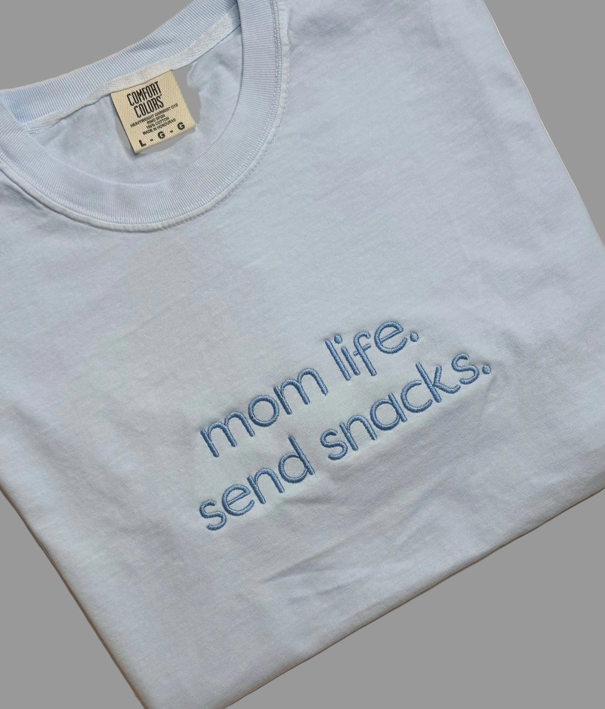 Light blue t-shirt with 'mom life. send snacks.' text on a gray background