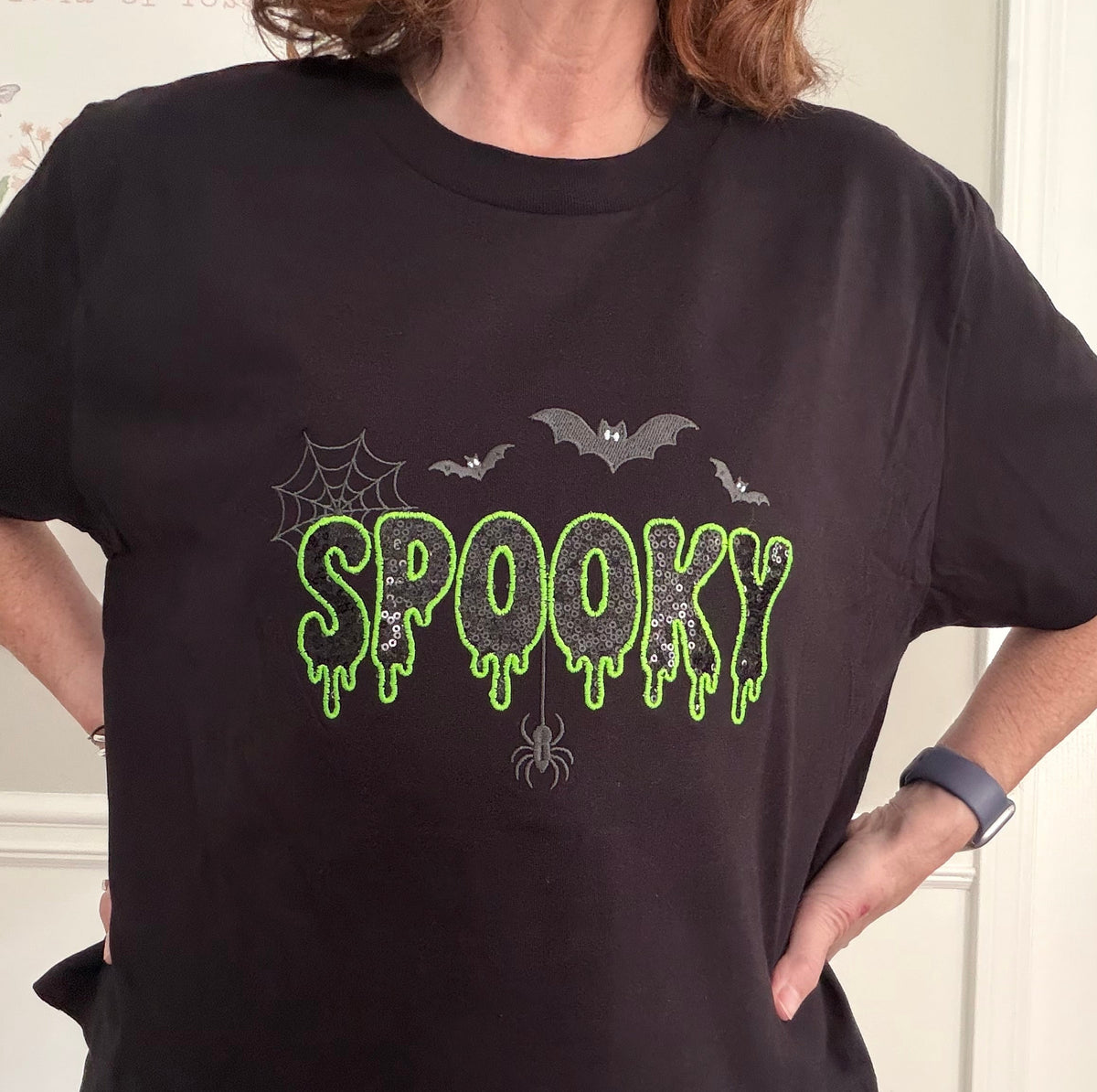 A person wearing a black short-sleeve T-shirt with a Halloween-themed graphic on the chest. The graphic features the word 'SPOOKY' in dripping neon green letters with a glittery black sequin inside, surrounded by dark gray spiderwebs, bats, and a spider.