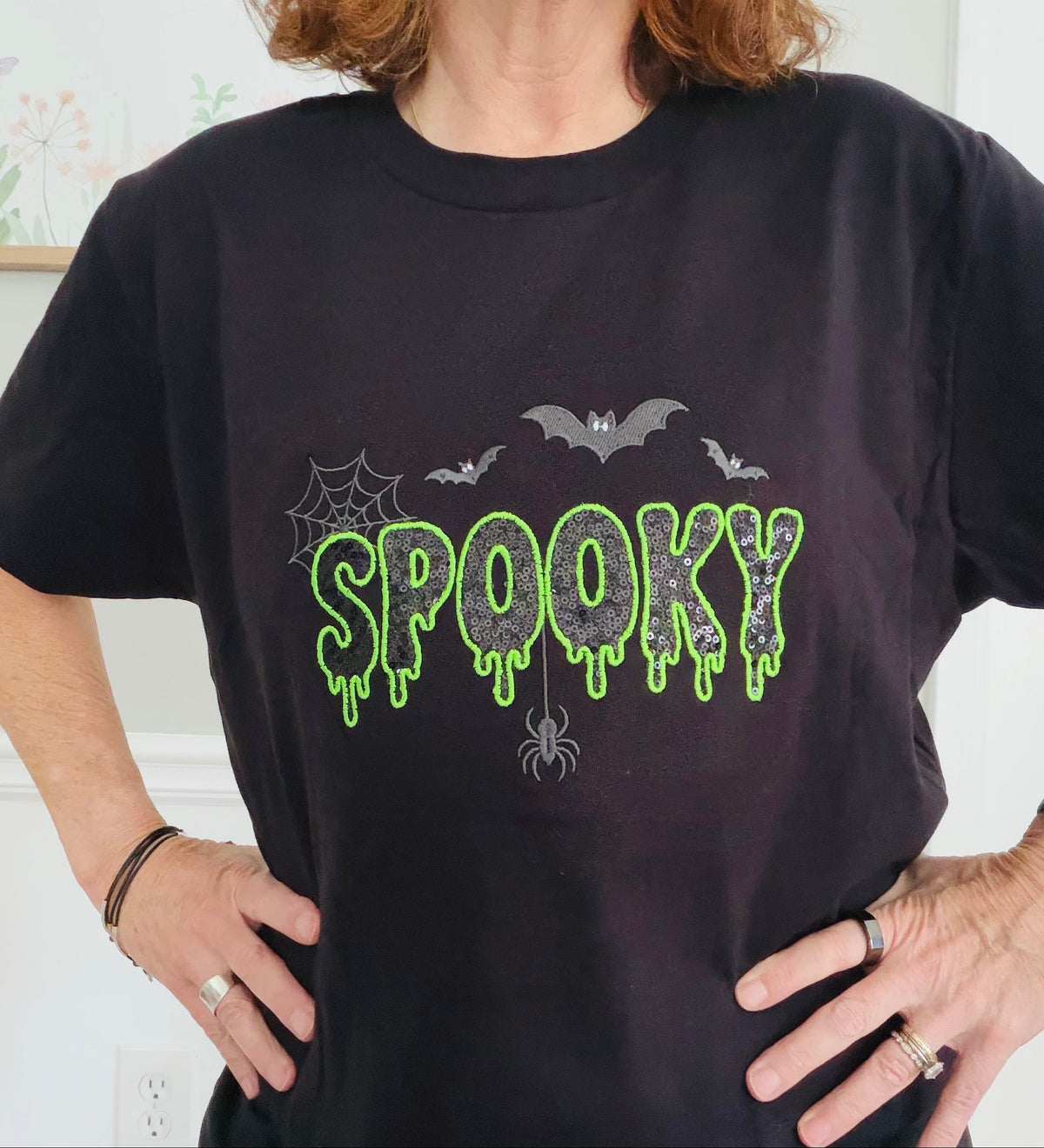 A person wearing a black short-sleeve T-shirt with a glittery 'SPOOKY' graphic in neon green and gray, featuring bats, a spider, and spiderwebs.