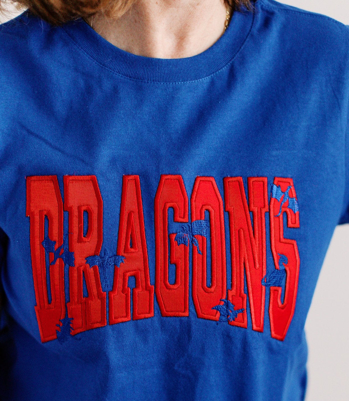 A close-up of a person wearing a royal blue t-shirt with a large red arched 'DRAGONS' graphic across the chest, featuring small blue dragon illustrations.