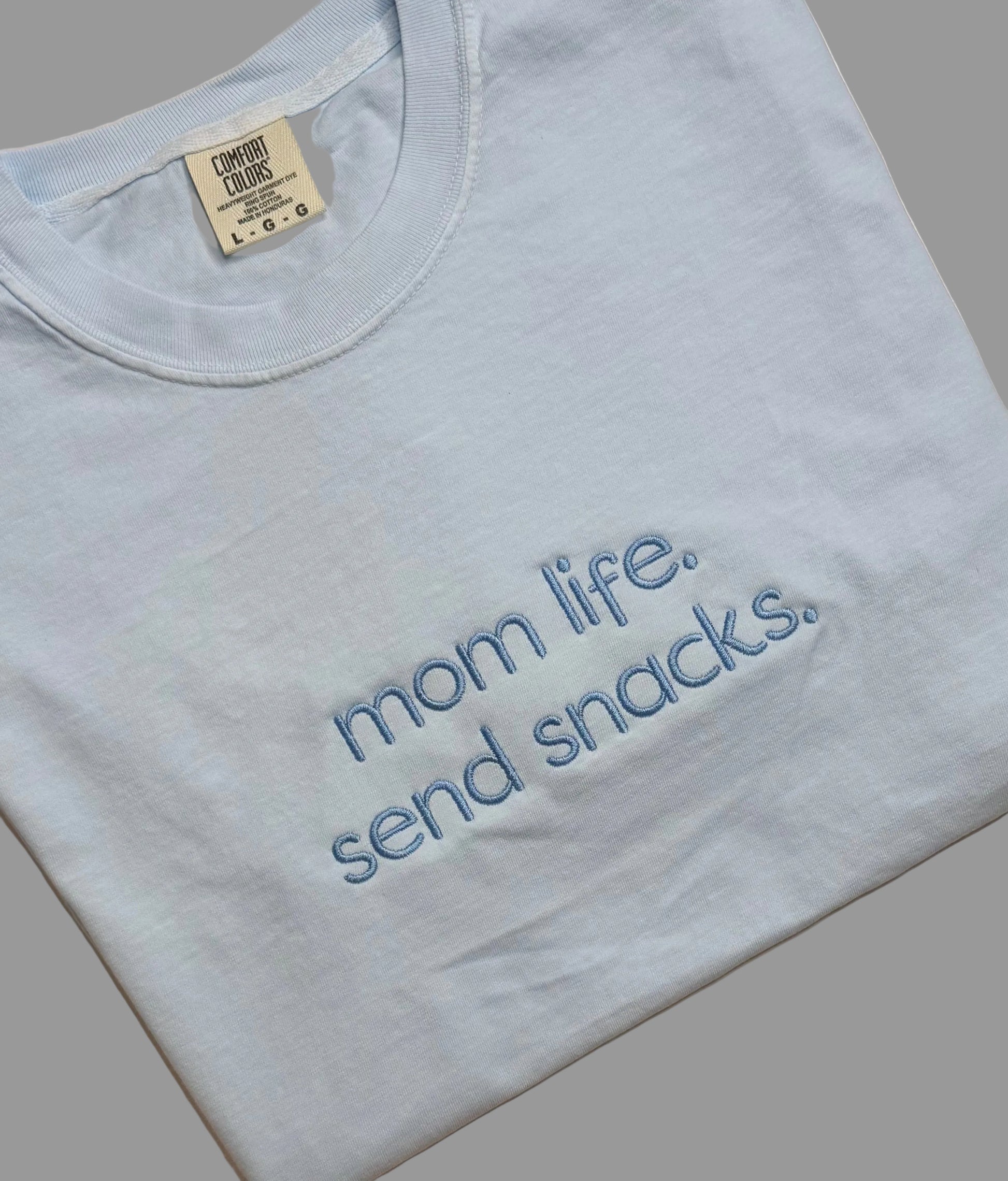 Light blue t-shirt with 'mom life. send snacks.' text on a gray background