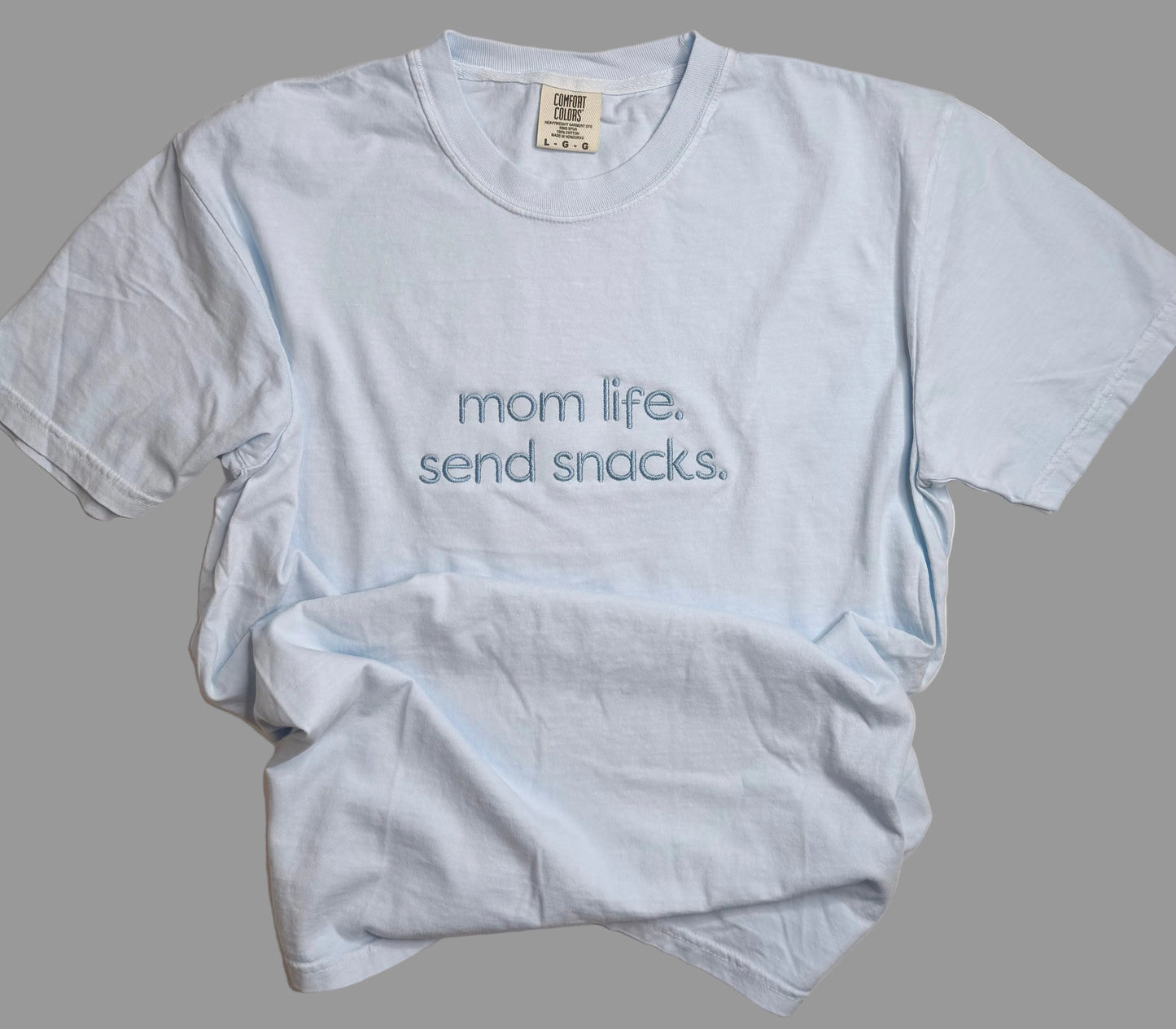Light blue t-shirt with 'mom life. send snacks.' text on a gray background