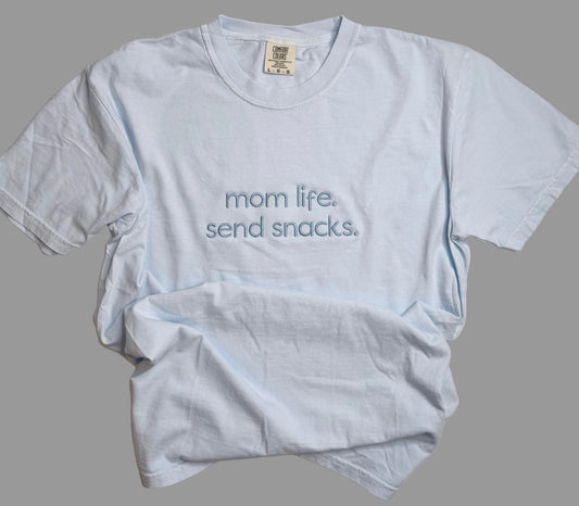 Light blue t-shirt with 'mom life. send snacks.' text on a gray background