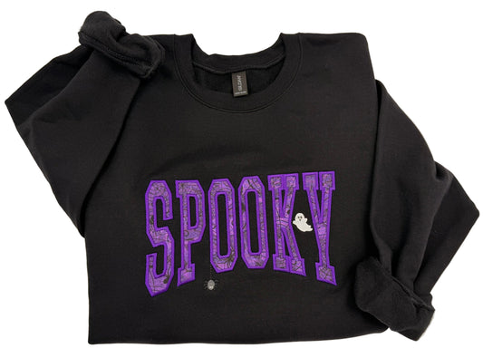 A black long-sleeve sweatshirt with 'SPOOKY' embroidery applique across the front in purple patterned letters, featuring a small white ghost detail, laid flat on a light beige background.