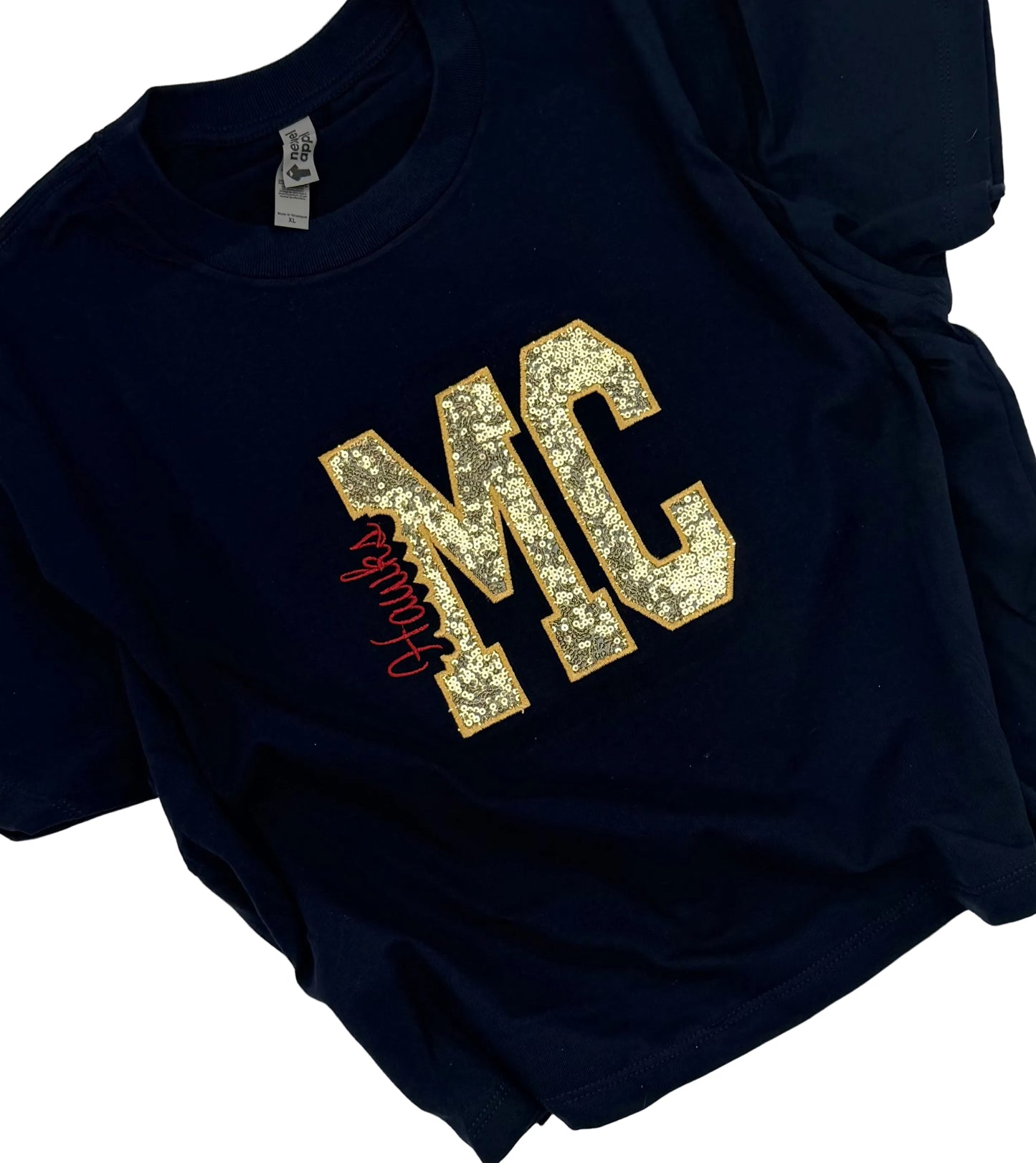 A navy blue short-sleeve t-shirt laid flat on a white background, featuring a large gold glitter 'MC' graphic on the chest with a red cursive script overlay.
