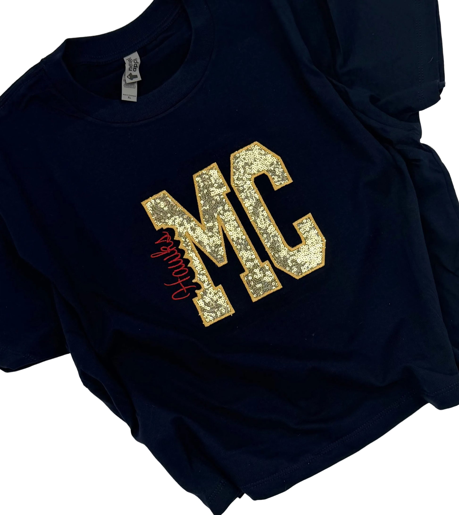 A navy blue short-sleeve t-shirt laid flat on a white background, featuring a large gold glitter 'MC' graphic on the chest with a red cursive script overlay.
