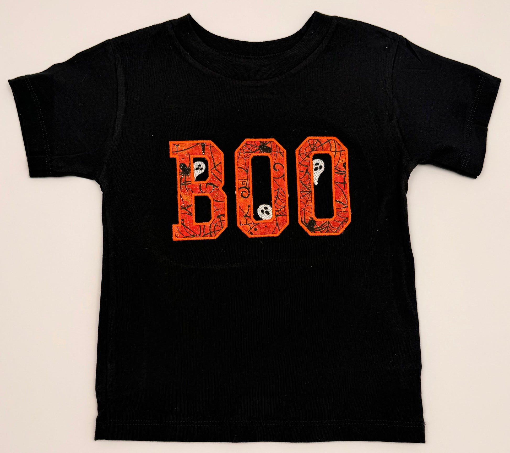 A black short-sleeve kids' t-shirt featuring the word 'BOO' printed across the chest in orange letters filled with spooky Halloween-themed illustrations like ghosts and bats, shown against a light beige background.